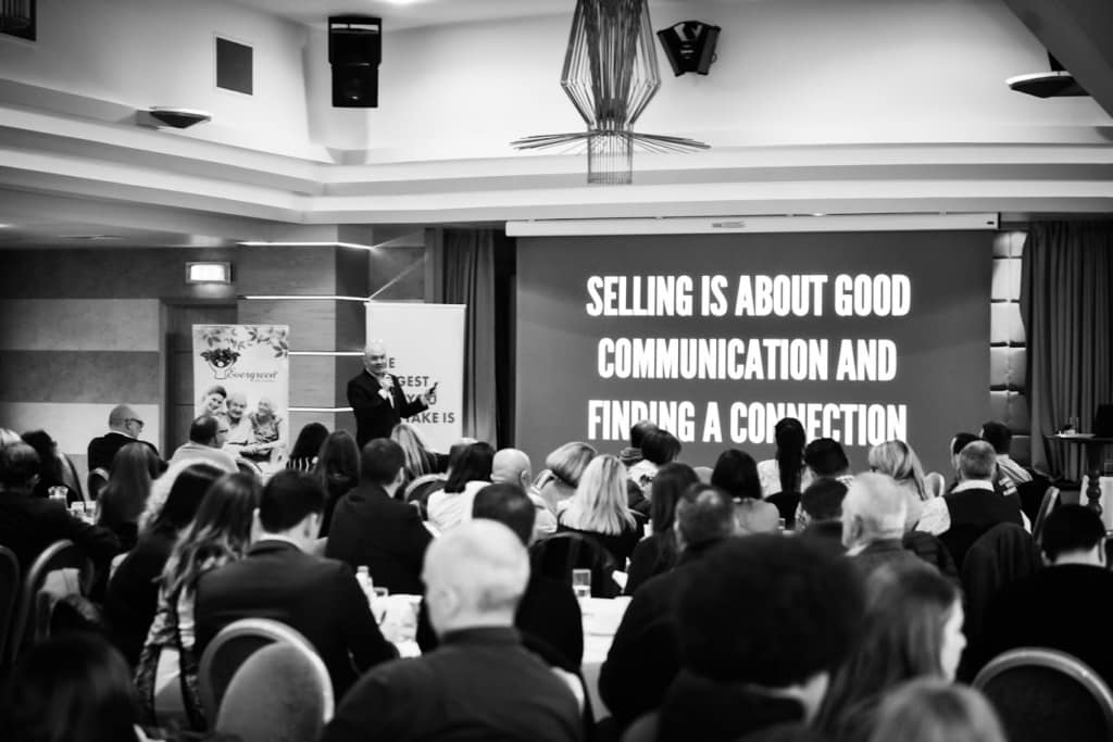 Speaker presenting to a seated audience in a conference hall with a large screen that reads selling is about good communication and finding a connection