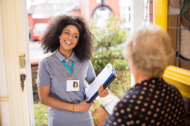 Young caregiver with a clipboard greets an elderly woman at the doorway smiling in a sunny entryway