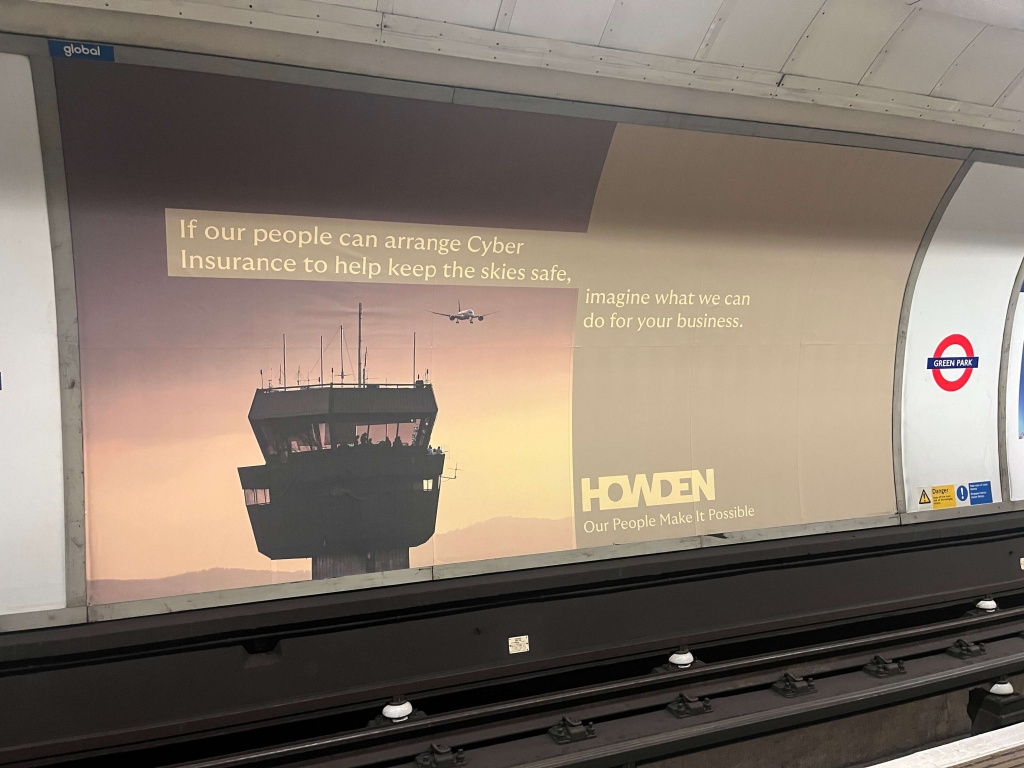 Subway platform advertisement showing a control tower and airplane with the cyber insurance slogan and a green park tube sign on the right