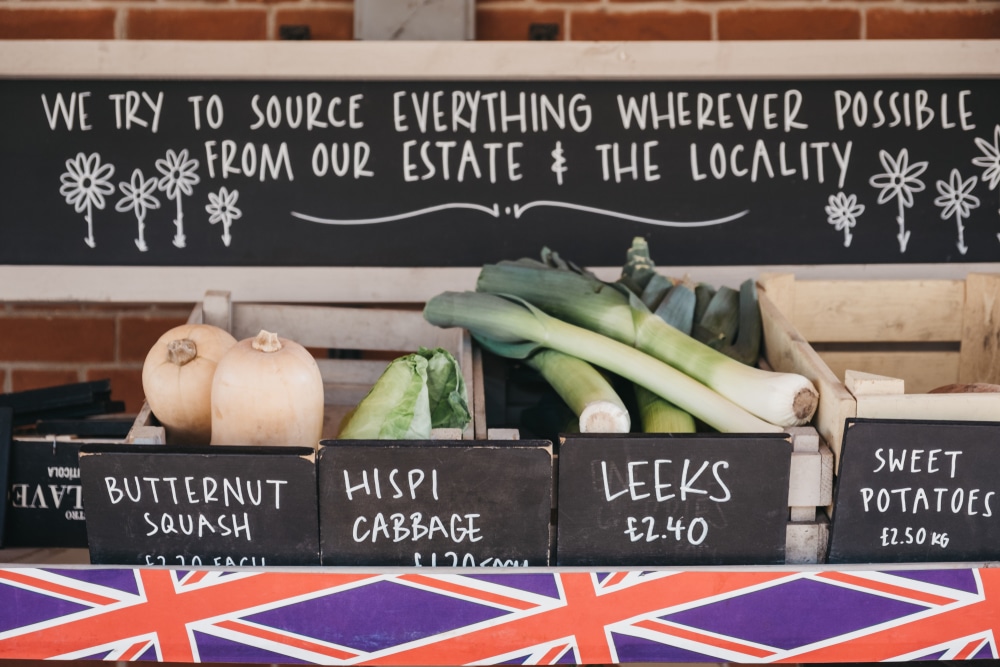 Benefits of sourcing local ingredients Market stall with crates of fresh vegetables labeled butternut squash hispi cabbage leeks and sweet potatoes at a local market A chalkboard signs about sourcing from estate and locality is above