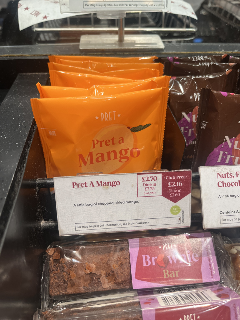 Img 7444 Front row of orange pret a mango snack bars on a store shelf beside brown chocolate bars