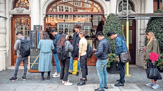 Screenshot 2024 11 18 at 13 12 35 A line of people standing outside a storefront with arched glass doors on a city street many carrying backpacks