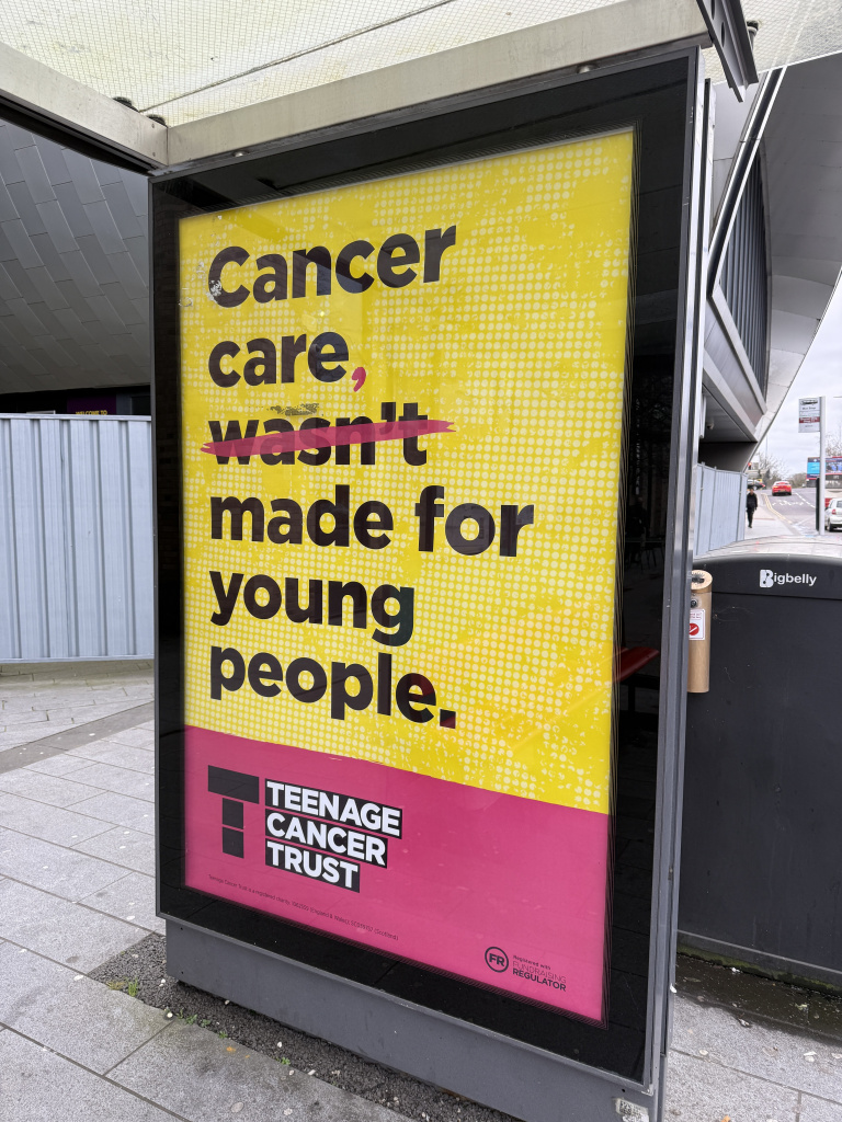 Bright yellow bus shelter poster that reads cancer care wasnt made for young people with a pink band for teenage cancer trust at the bottom
