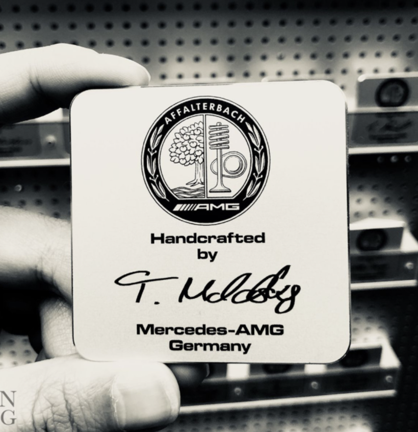 Close up of a square black and white mercedes amg badge tag held by fingers reading handcrafted by with a signature and germany