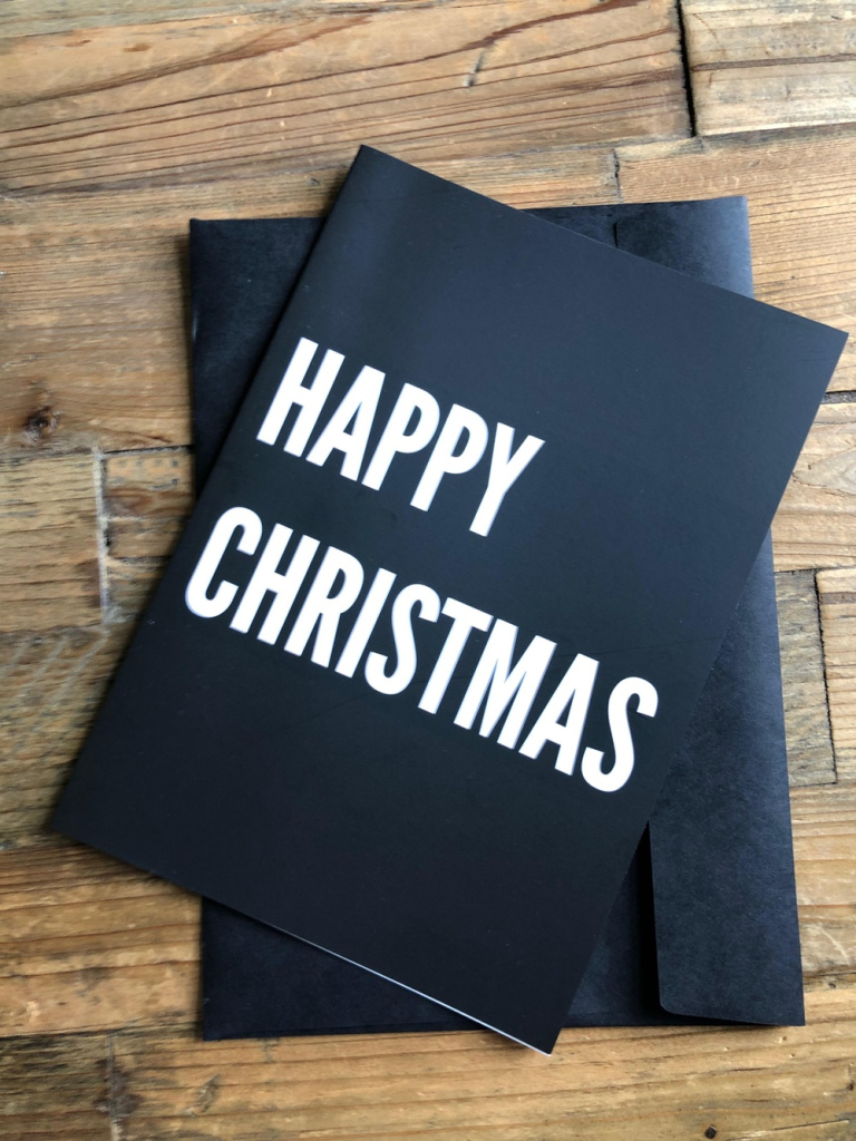 Black greeting card with bold white happy christmas text on a wooden surface resting on a dark envelope beneath it