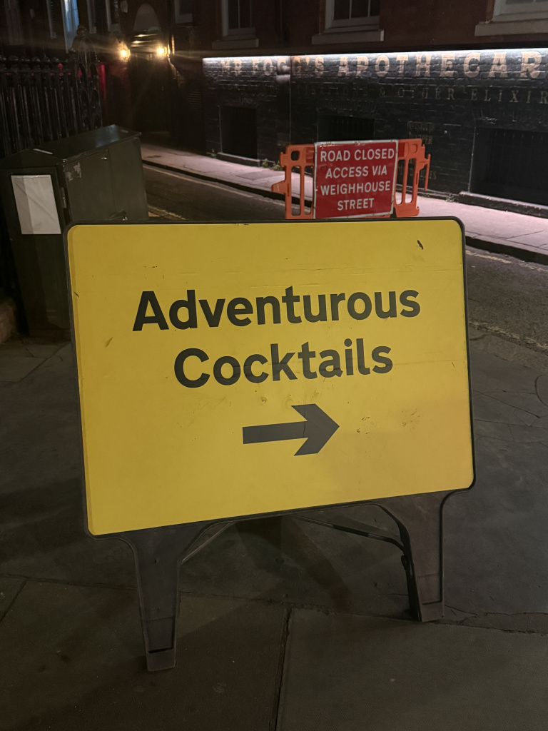 Yellow sign reading adventurous cocktails with an arrow pointing right street scene behind it and a red road closed barrier nearby nighttime