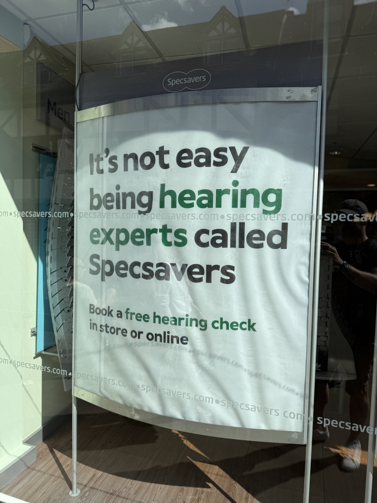 Advertising poster in a shop window reading its not easy being hearing experts called specsavers with the specsavers logo visible on top portion