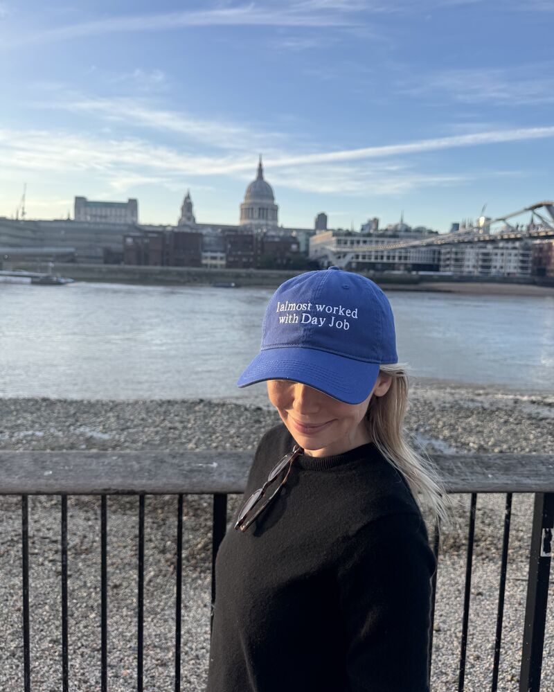 Woman in a black sweater stands by a railing along a river wearing a blue cap that reads i almost worked with day job with sunglasses hanging from her neckline and a city skyline in the background