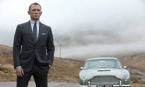 Well dressed man in a dark suit standing with hands in pockets beside a vintage light colored car on a misty hillside road with fog in the background