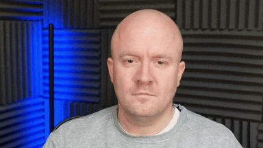 Bald man in a gray sweatshirt with a neutral expression seated in a studio with black acoustic panels and blue lighting behind him