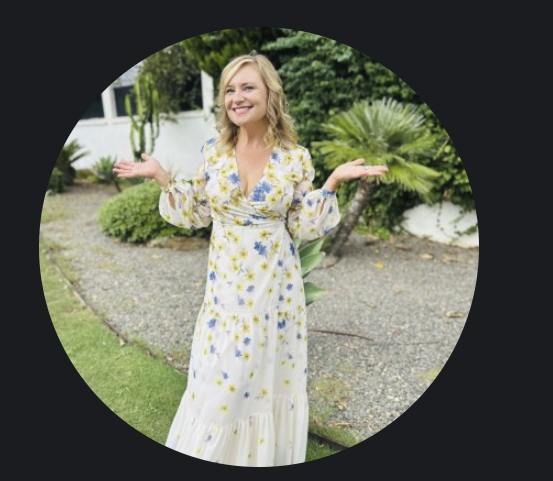 Smiling woman in a pale yellow floral dress stands outdoors with arms raised