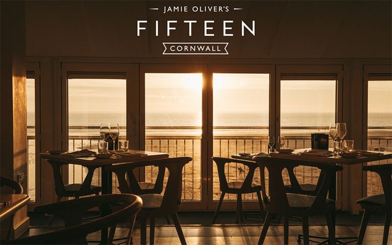 Jamie oliver's Fifteen Cornwall restaurant interior at sunset, with tables set near windows overlooking the ocean.