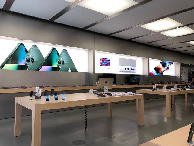 Images Bright electronics showroom with wooden tables displaying iphones and ipads and large back wall iphone graphics behind them Edge note avoid question mark
