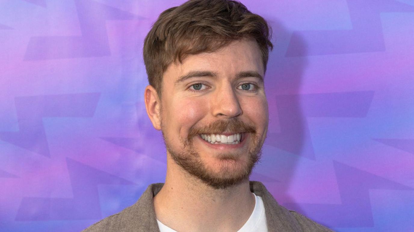 Smiling young man with short brown hair and a beard wearing a light jacket against a purple geometric backdrop