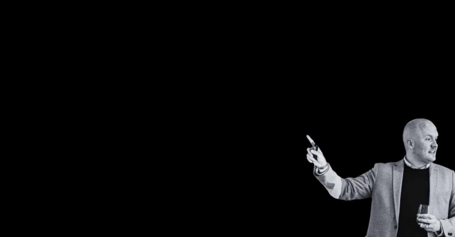 A man in a blazer points to the left while holding a glass set against a dark background as if giving a presentation