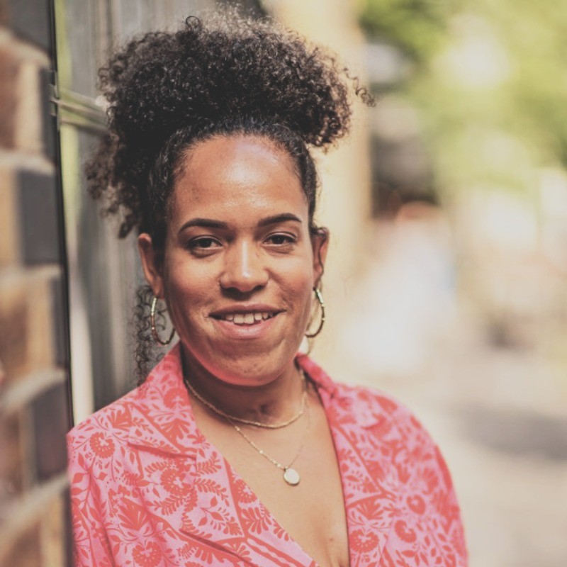 Woman with curly hair leaning against a brick wall smiling at the camera in a pink patterned top